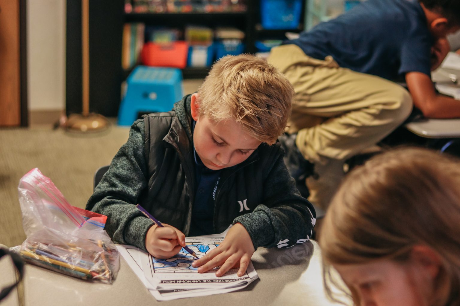 A boy coloring in an art assignment while in class as a teacher helps a student behind him