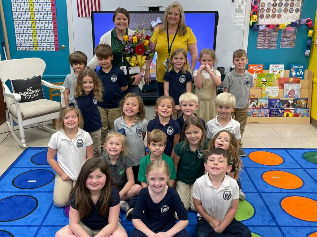 Teacher Megan Patterson smiling holding a bouquet of flowers in her classroom, with her whole class of students in-front of her